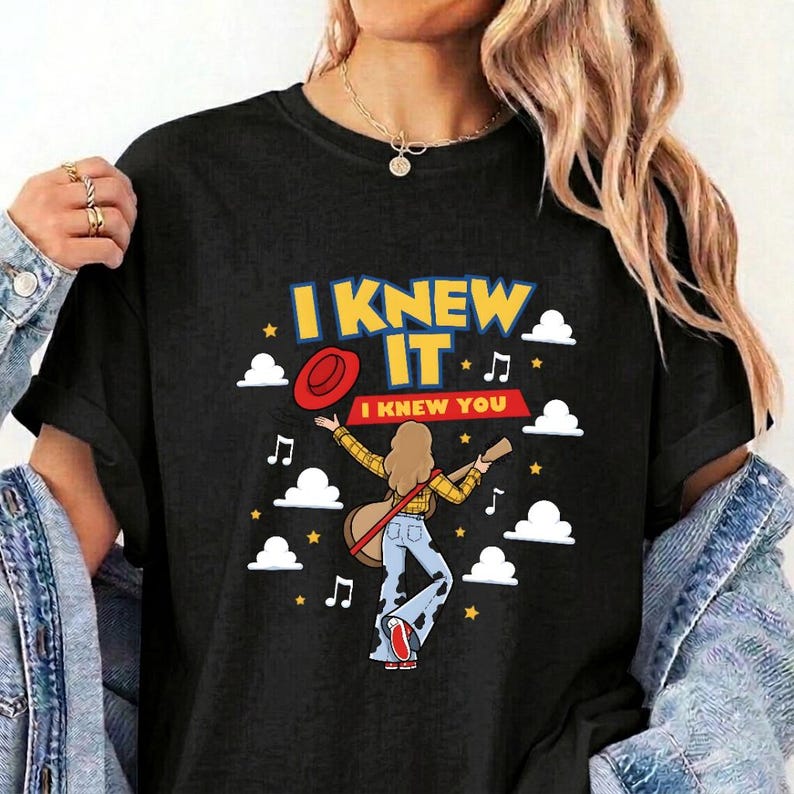 May include: A black t-shirt featuring a graphic of a person with long hair wearing a yellow plaid shirt and blue jeans, holding a guitar. The design includes a red hat, white clouds, musical notes, and the text I KNEW IT I KNEW YOU.