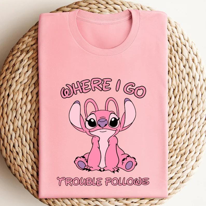May include: Pink t-shirt featuring a cartoon character with large ears and the text "WHERE I GO TROUBLE FOLLOWS" in a playful font. The design is centered on the shirt, which is folded on a woven surface.