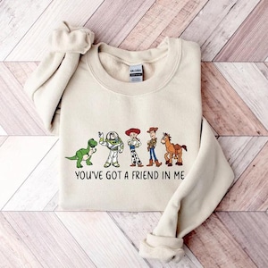 May include: A cream-colored sweatshirt featuring a graphic of cartoon characters and the text "YOU'VE GOT A FRIEND IN ME". The design includes a green dinosaur, a space ranger, a cowgirl, a cowboy, and a brown horse.