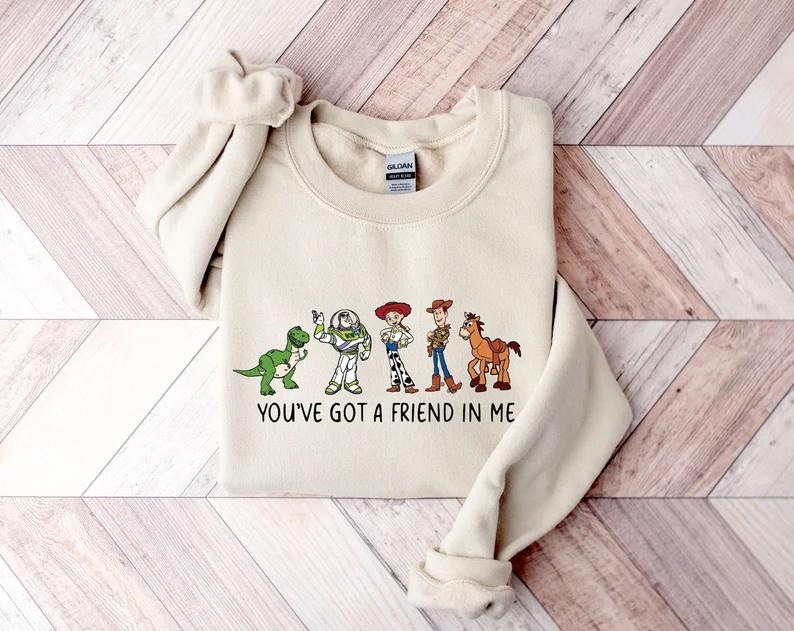 May include: A cream-colored sweatshirt featuring a graphic of cartoon characters and the text "YOU'VE GOT A FRIEND IN ME". The design includes a green dinosaur, a space ranger, a cowgirl, a cowboy, and a brown horse.