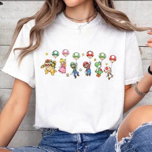 May include: A white short-sleeve t-shirt featuring a graphic print of various video game characters including Bowser, Princess Peach, Luigi, Mario, Yoshi, and Toad, each holding a colorful mushroom-shaped balloon against a plain white background.