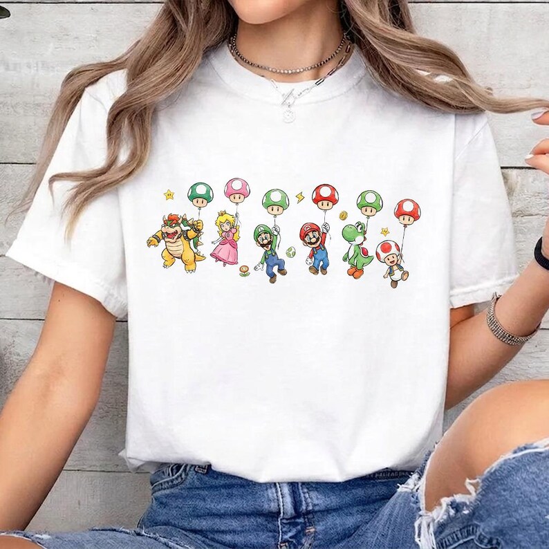 May include: A white short-sleeve t-shirt featuring a graphic print of various video game characters including Bowser, Princess Peach, Luigi, Mario, Yoshi, and Toad, each holding a colorful mushroom-shaped balloon against a plain white background.
