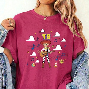 May include: A dark pink t-shirt featuring a graphic of a cartoon cowgirl playing an acoustic guitar. The design includes the text TS, white clouds, blue musical notes, a yellow ball, a traffic cone, and a yellow sheriff star on a dark pink background.