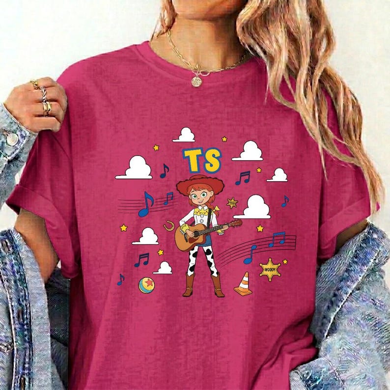 May include: A dark pink t-shirt featuring a graphic of a cartoon cowgirl playing an acoustic guitar. The design includes the text TS, white clouds, blue musical notes, a yellow ball, a traffic cone, and a yellow sheriff star on a dark pink background.