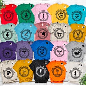 Peut inclure: Une collection de vingt t-shirts colorés, chacun avec un design unique dans un cadre circulaire. Les chemises affichent le texte "CAMP HALF-BLOOD" et divers symboles et noms de la mythologie grecque, dont "Déméter" et "Apollon".