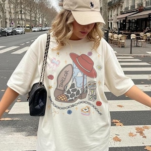 May include: An off-white short-sleeve t-shirt featuring a graphic print of a cowboy boot with a cow-print pattern, a red hat, the name Taylor, the number 13, and stacked blocks with the letters T, S, and S, surrounded by small floral and bow icons.