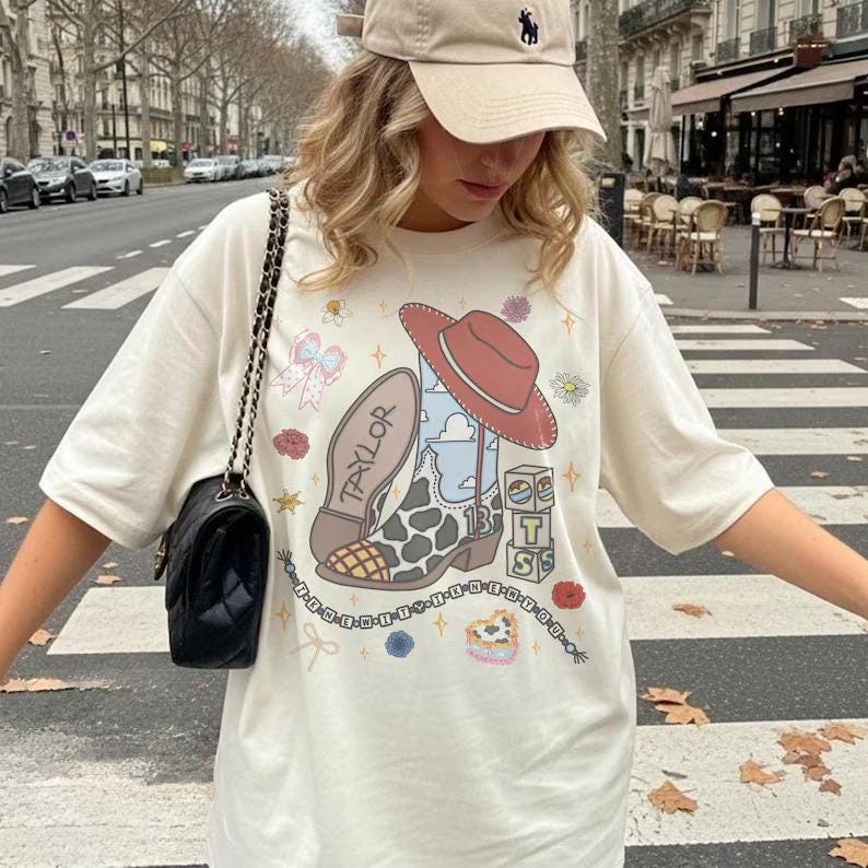May include: An off-white short-sleeve t-shirt featuring a graphic print of a cowboy boot with a cow-print pattern, a red hat, the name Taylor, the number 13, and stacked blocks with the letters T, S, and S, surrounded by small floral and bow icons.
