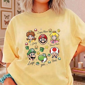 May include: A pale yellow short-sleeve t-shirt featuring a colorful graphic print of Super Mario characters including Mario, Luigi, Princess Peach, Daisy, Yoshi, and Toad, along with game icons and the text Super Mario, Daisy, Princess Peach, Luigi, Yoshi, and Toad.