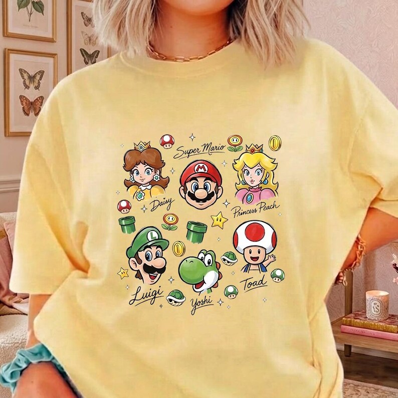 May include: A pale yellow short-sleeve t-shirt featuring a colorful graphic print of Super Mario characters including Mario, Luigi, Princess Peach, Daisy, Yoshi, and Toad, along with game icons and the text Super Mario, Daisy, Princess Peach, Luigi, Yoshi, and Toad.