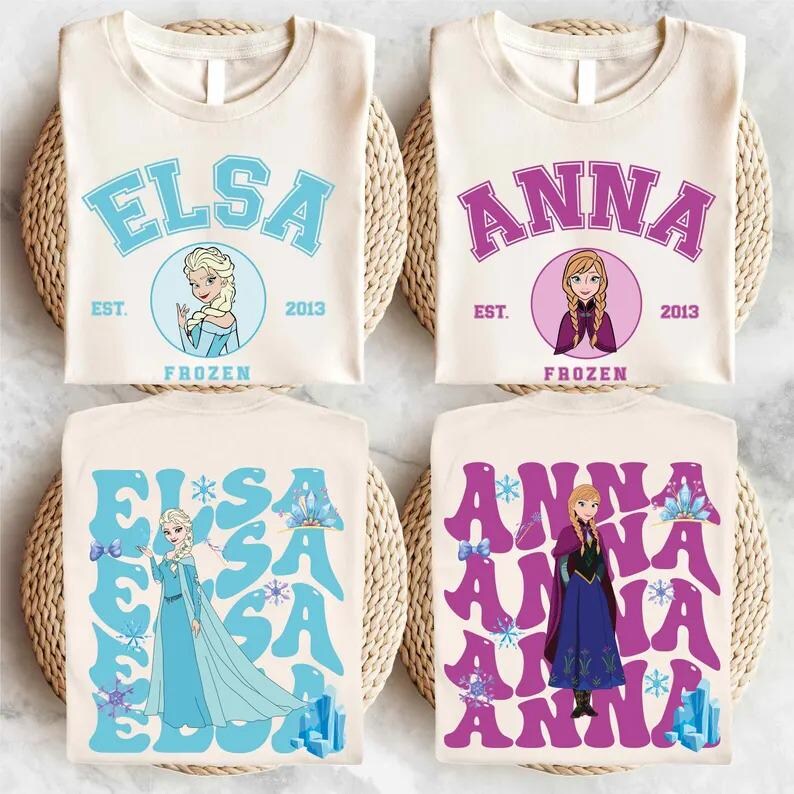 May include: Four cream-colored t-shirts featuring designs from the movie "Frozen." Two shirts display "ELSA" and "ANNA" in blue and purple, with character illustrations. The other two shirts have the names in a stylized font.