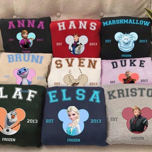 May include: A collection of sweatshirts in various colors, each featuring a character from the movie "Frozen". The sweatshirts display character names like Anna, Hans, and Olaf, along with the year "2013" and the word "Frozen".
