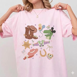 May include: A light pink t-shirt featuring a colorful graphic design. The design includes cartoon illustrations of a cowboy hat, boots, a sheriff badge, a dinosaur, and other playful elements. The shirt has a relaxed fit.