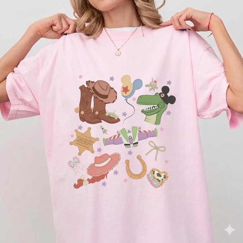 May include: A light pink t-shirt featuring a colorful graphic design. The design includes cartoon illustrations of a cowboy hat, boots, a sheriff badge, a dinosaur, and other playful elements. The shirt has a relaxed fit.