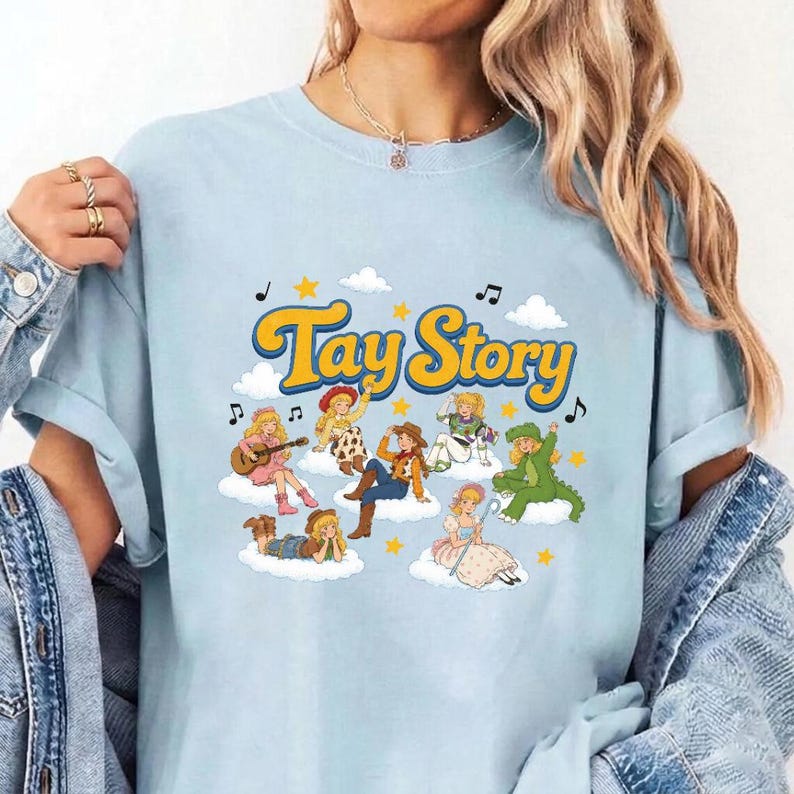 Pu&ograve; includere: Una t-shirt azzurra con una stampa grafica che riporta il testo Tay Story in lettere gialle. Il design include diversi personaggi illustrati con capelli biondi seduti su nuvole bianche, circondati da stelle gialle e note musicali nere.
