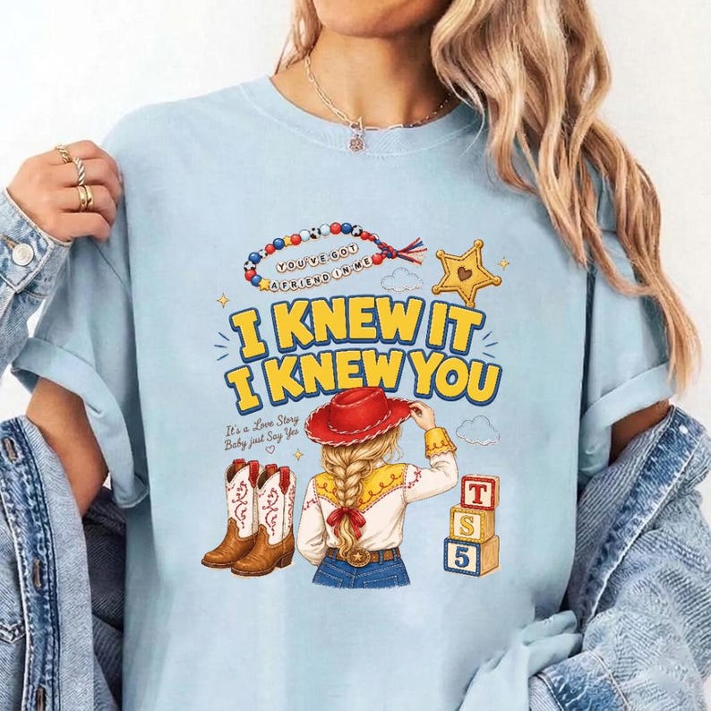 May include: A light blue t-shirt featuring a graphic print with the text I KNEW IT I KNEW YOU, a beaded necklace, a yellow star, a red cowboy hat, blonde braided hair, brown cowboy boots, and three stacked blocks with the letters T, S, and 5.