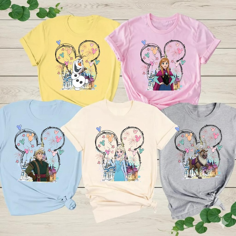 May include: Five short-sleeved t-shirts in yellow, pink, light blue, cream, and grey. Each shirt has a Disney-themed graphic with characters and fireworks inside a Mickey Mouse head outline.