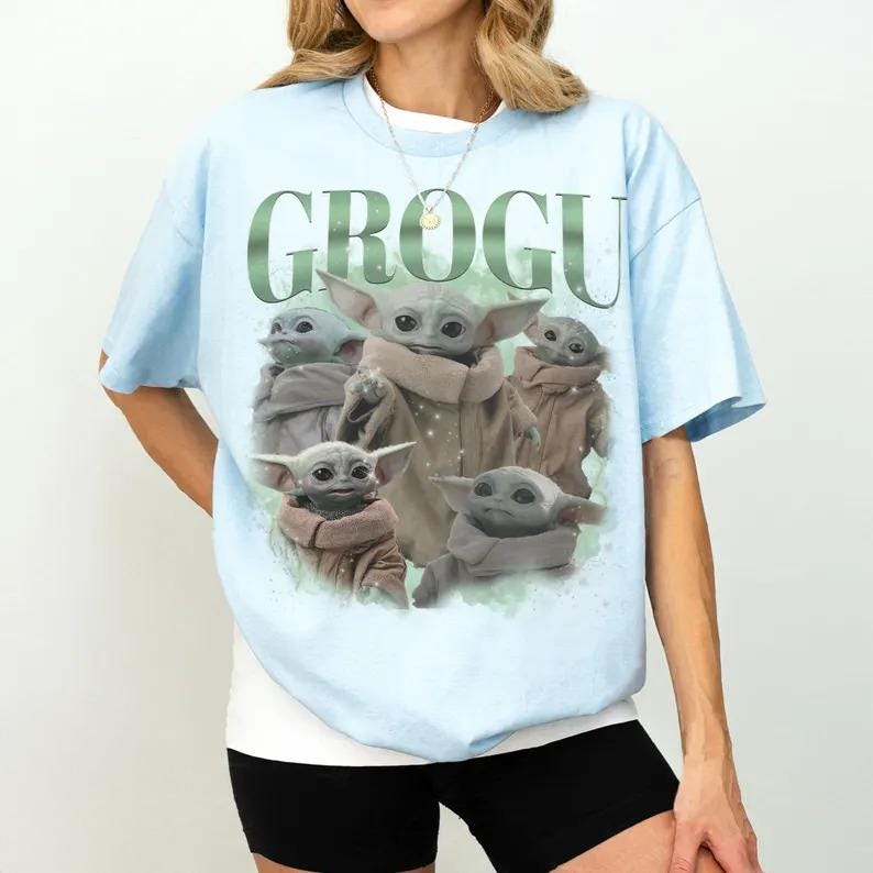 Może przedstawiać: Jasnoniebieski t-shirt z nadrukiem graficznym przedstawiającym kilka postaci Grogu w r&oacute;żnych pozach. Napis GROGU jest wydrukowany dużymi zielonymi literami nad postaciami. Wz&oacute;r zawiera subtelne akcenty w kształcie gwiazdek oraz delikatne zielone tło.