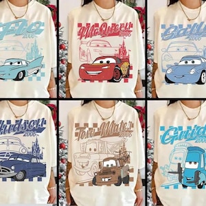 Op de afbeelding: Cr&egrave;mekleurige T-shirts met verschillende personages uit de animatiefilm "Cars". Elk shirt toont de naam van een ander personage, het jaar 2006 en een illustratie van de auto van het personage in blauw, rood en bruin.