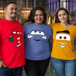 May include: Three people wearing t-shirts with cartoon car faces. The shirts are red, blue, and yellow. The red shirt has a car face with a green and white design. The blue shirt has a blue car face. The yellow shirt has a car face with a black and white design.