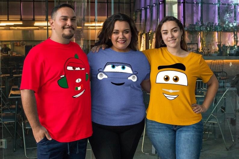 May include: Three people wearing t-shirts with cartoon car faces. The shirts are red, blue, and yellow. The red shirt has a car face with a green and white design. The blue shirt has a blue car face. The yellow shirt has a car face with a black and white design.