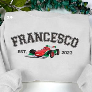 May include: White sweatshirt featuring the name "FRANCESCO" in a black, collegiate font. A red, green, and white race car graphic is below the name. The text "EST. 2023" is below the car. The sweatshirt has a crew neck and long sleeves.