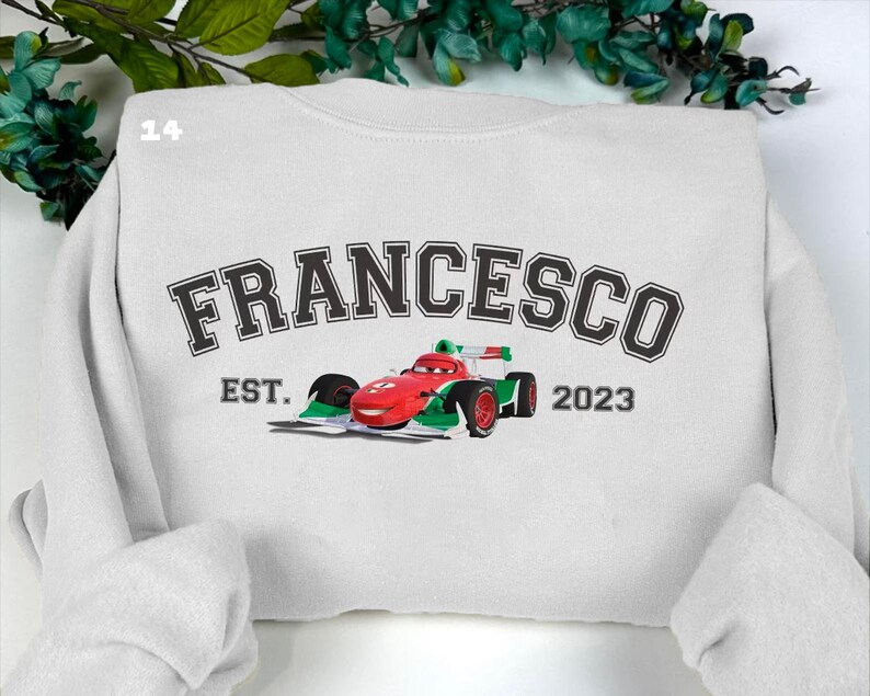 May include: White sweatshirt featuring the name "FRANCESCO" in a black, collegiate font. A red, green, and white race car graphic is below the name. The text "EST. 2023" is below the car. The sweatshirt has a crew neck and long sleeves.