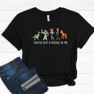 May include: Black t-shirt featuring a graphic of cartoon characters from the movie Toy Story. The characters include a green dinosaur, Buzz Lightyear, Jessie, Woody, and a horse. The text "YOU'VE GOT A FRIEND IN ME" is printed below the characters.