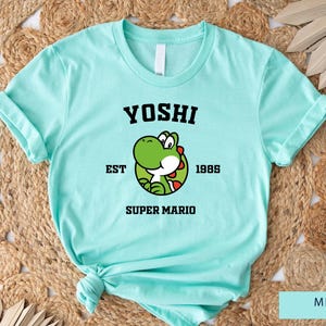 May include: Mint green t-shirt with a black graphic of Yoshi from Super Mario Bros. The graphic includes the text "Yoshi" in a collegiate font, "EST 1985", and "Super Mario".