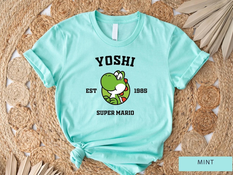 May include: Mint green t-shirt with a black graphic of Yoshi from Super Mario Bros. The graphic includes the text "Yoshi" in a collegiate font, "EST 1985", and "Super Mario".