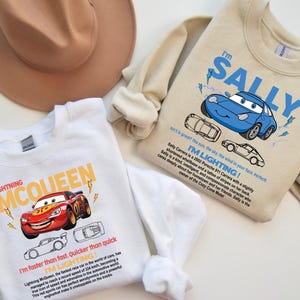 May include: Two sweatshirts featuring Cars movie characters. One is white with Lightning McQueen graphics and text. The other is beige with Sally Carrera graphics and text. A brown hat is in the background.