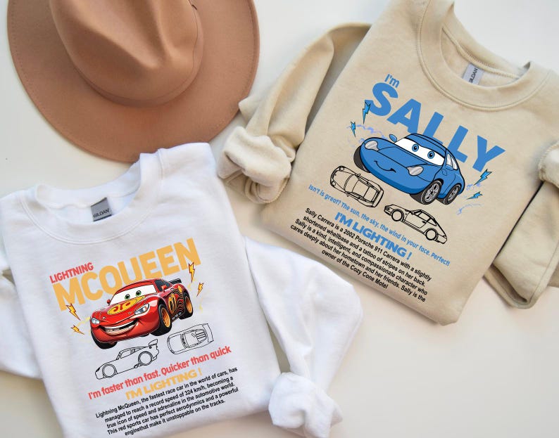 May include: Two sweatshirts featuring Cars movie characters. One is white with Lightning McQueen graphics and text. The other is beige with Sally Carrera graphics and text. A brown hat is in the background.