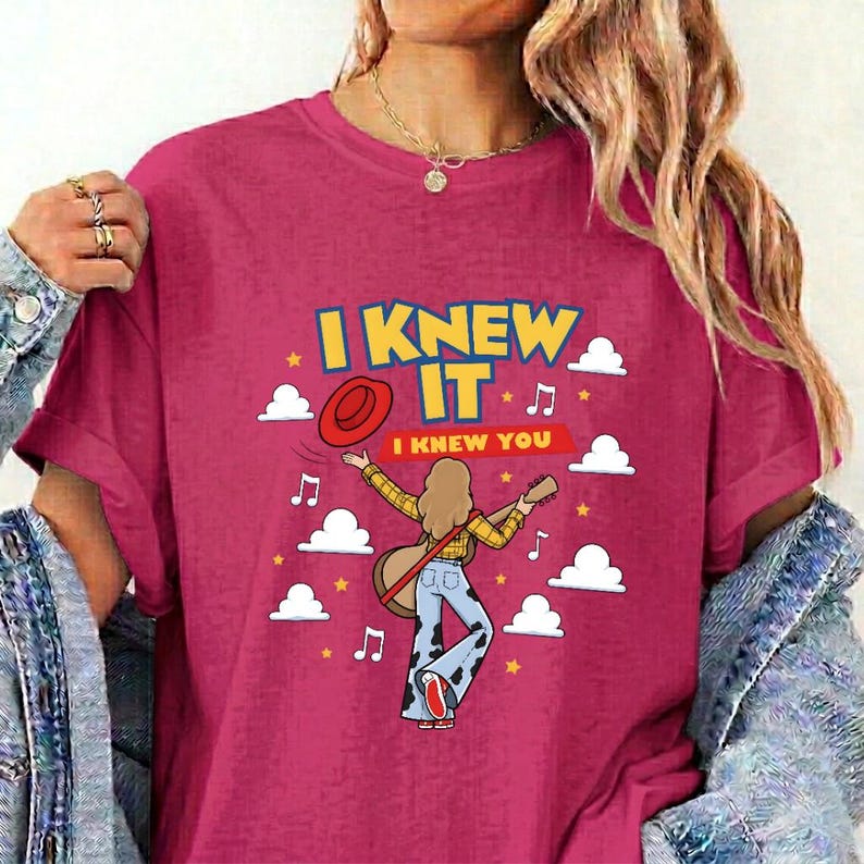 May include: A magenta t-shirt featuring a graphic of a person with long hair wearing a yellow plaid shirt and cow-print pants, holding a guitar. The text reads I KNEW IT and I KNEW YOU, surrounded by white clouds, yellow stars, and musical notes.