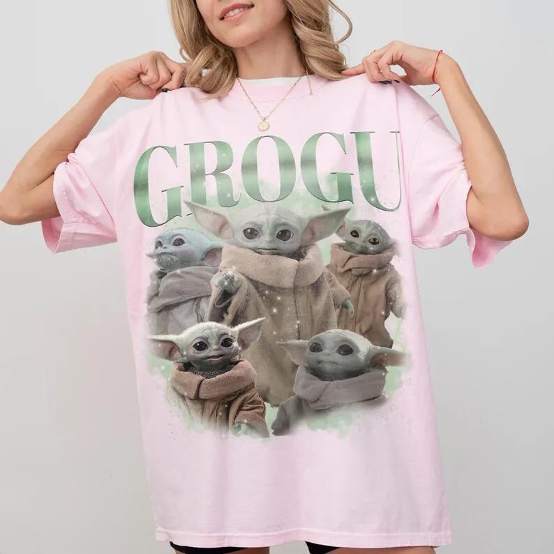 Może przedstawiać: Jasnor&oacute;żowy t-shirt z kr&oacute;tkim rękawem z dużym nadrukiem graficznym przedstawiającym pięć postaci Grogu w r&oacute;żnych pozach. Napis GROGU jest wydrukowany dużymi, pogrubionymi, zielonymi literami w g&oacute;rnej części grafiki. Wz&oacute;r zawiera subtelne białe iskierki.