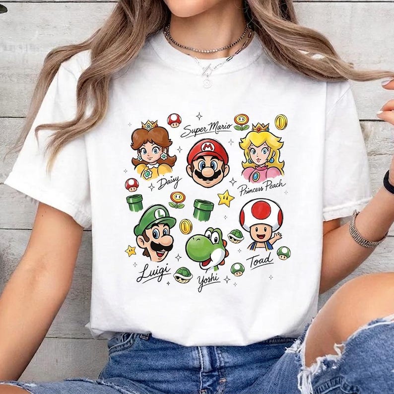 May include: A white short-sleeve t-shirt featuring a colorful graphic print of Super Mario characters including Mario, Luigi, Princess Peach, Daisy, Yoshi, and Toad, along with game icons and the names of the characters written in black script.