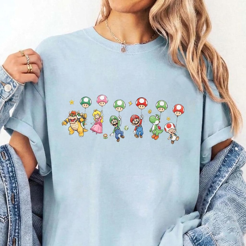 May include: A light blue t-shirt featuring a graphic print of various Nintendo characters including Bowser, Princess Peach, Luigi, Mario, Yoshi, and Toad, each holding a colorful mushroom-shaped balloon against a plain background.