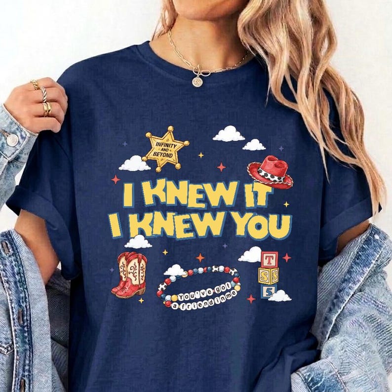 Puede incluir: Una camiseta azul marino con un estampado gr&aacute;fico que incluye el texto I KNEW IT I KNEW YOU. El dise&ntilde;o presenta una estrella de sheriff amarilla, un sombrero de vaquero rojo, botas vaqueras rojas, una pulsera de cuentas y bloques de letras.