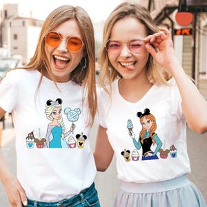 May include: Two women wearing white t-shirts with cartoon character graphics. One shirt features Elsa with cotton candy, the other Anna with ice cream. Both shirts have Mickey Mouse ear headbands and colorful dessert designs.