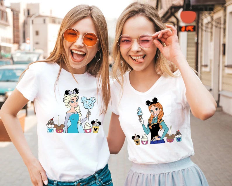 May include: Two women wearing white t-shirts with cartoon character graphics. One shirt features Elsa with cotton candy, the other Anna with ice cream. Both shirts have Mickey Mouse ear headbands and colorful dessert designs.