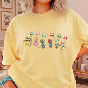 May include: A pale yellow t-shirt featuring a graphic print of Bowser, Princess Peach, Luigi, Mario, Yoshi, and Toad. Each character is holding a string attached to a floating mushroom-shaped balloon. The design includes small yellow stars.