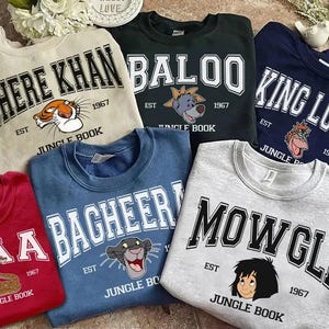 May include: A collection of six crewneck sweatshirts in red, beige, black, blue, navy, and grey. Each features a character from The Jungle Book, including Kaa, Shere Khan, Baloo, Bagheera, King Louie, and Mowgli, with their names and the text Jungle Book 1967.