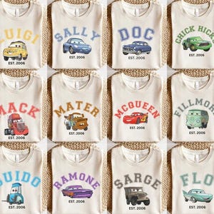 May include: A selection of beige t-shirts, each adorned with a cartoon car character's name in a collegiate style. The shirts also feature an image of the car and the text "EST. 2006". Characters include Luigi, Sally, Doc, and Chick Hicks, among others.