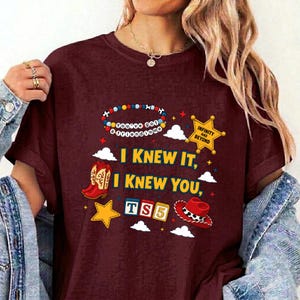 May include: A maroon t-shirt featuring a graphic print with a colorful bead necklace, a yellow sheriff star with the text INFINITY AND BEYOND, red cowboy boots, alphabet blocks spelling TS5, and a red cowboy hat, with the text I KNEW IT, I KNEW YOU.
