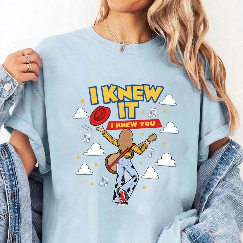 May include: A light blue t-shirt featuring a graphic of a person with long hair playing a guitar, wearing a yellow plaid shirt and cow-print pants. The text reads I KNEW IT and I KNEW YOU, accompanied by a red hat, musical notes, and white clouds.