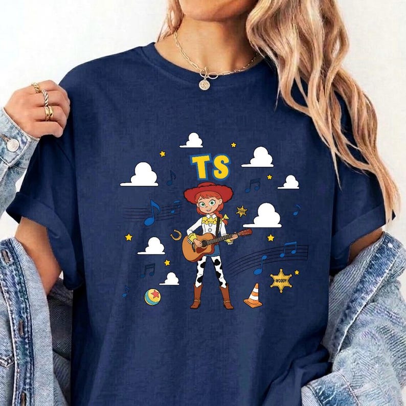 May include: A navy blue t-shirt featuring a graphic of a cartoon cowgirl playing an acoustic guitar. The design includes the yellow text TS, white clouds, blue musical notes, yellow stars, a colorful ball, a yellow sheriff star, and an orange cone.