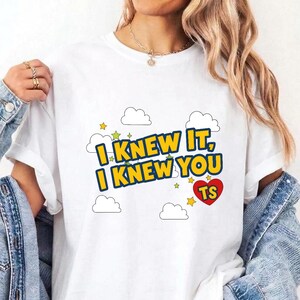 May include: A white short-sleeved t-shirt featuring a graphic print with the text I KNEW IT, I KNEW YOU in bold yellow letters with blue outlines. The design includes white cloud illustrations, small green and yellow stars, and a red heart with TS.
