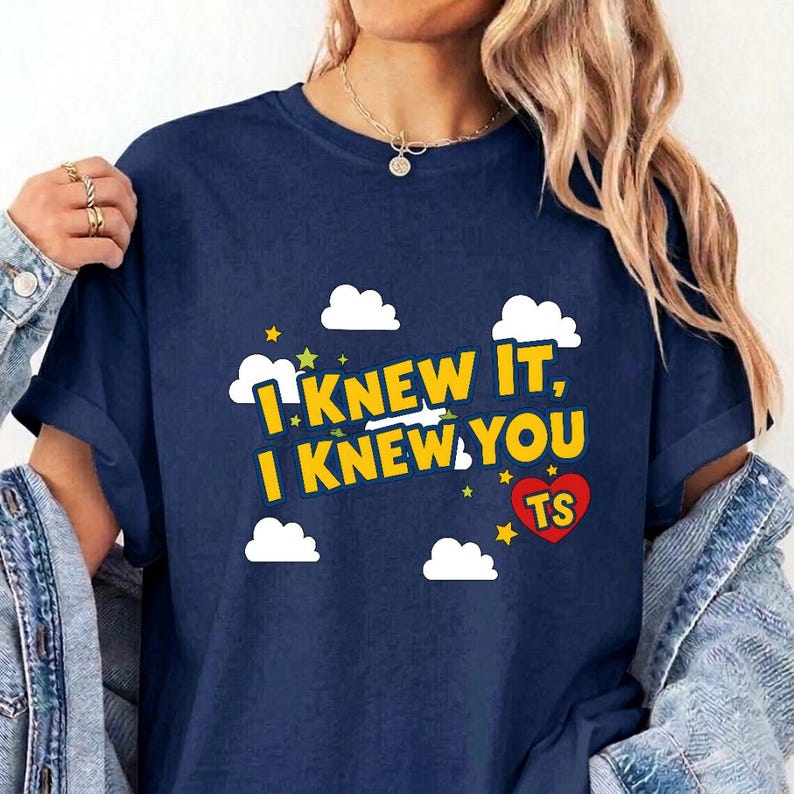 May include: A navy blue short-sleeved t-shirt featuring a graphic print with white clouds, yellow stars, and the yellow text "I KNEW IT, I KNEW YOU" alongside a red heart containing the letters "TS" in yellow, set against a solid dark blue background.