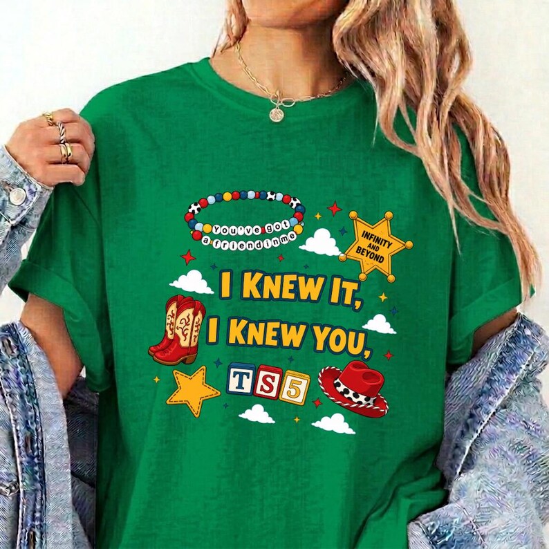 May include: A green t-shirt featuring a graphic print with a colorful beaded bracelet, a yellow sheriff star, red cowboy boots, a red cowboy hat, and blocks spelling TS5. The text reads I KNEW IT, I KNEW YOU, and INFINITY AND BEYOND.
