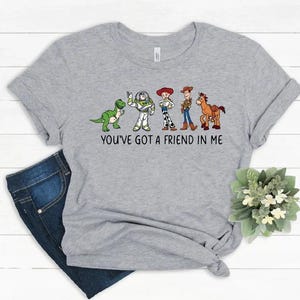 May include: A light gray t-shirt featuring a graphic of cartoon characters from the movie Toy Story. The characters include a green dinosaur, Buzz Lightyear, Jessie, Woody, and a horse. The text "YOU'VE GOT A FRIEND IN ME" is printed below the characters.