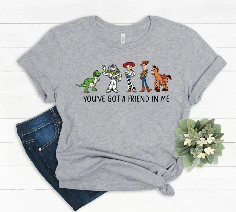 May include: A light gray t-shirt featuring a graphic of cartoon characters from the movie Toy Story. The characters include a green dinosaur, Buzz Lightyear, Jessie, Woody, and a horse. The text "YOU'VE GOT A FRIEND IN ME" is printed below the characters.