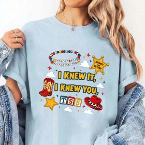 May include: A light blue t-shirt featuring a graphic print with the text I KNEW IT, I KNEW YOU, TS5, and INFINITY AND BEYOND. The design includes a colorful beaded bracelet, a yellow sheriff star, red cowboy boots, a red cowboy hat, and clouds.